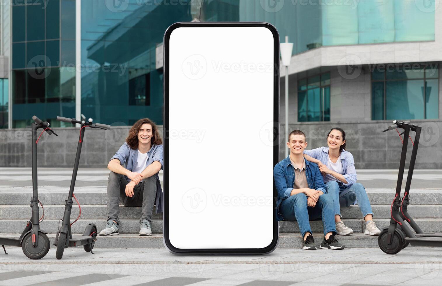 Three young adults are seated on steps near electric scooters and huge phone with blank screen in a modern urban environment. They are smiling and relaxed, embodying a carefree lifestyle photo