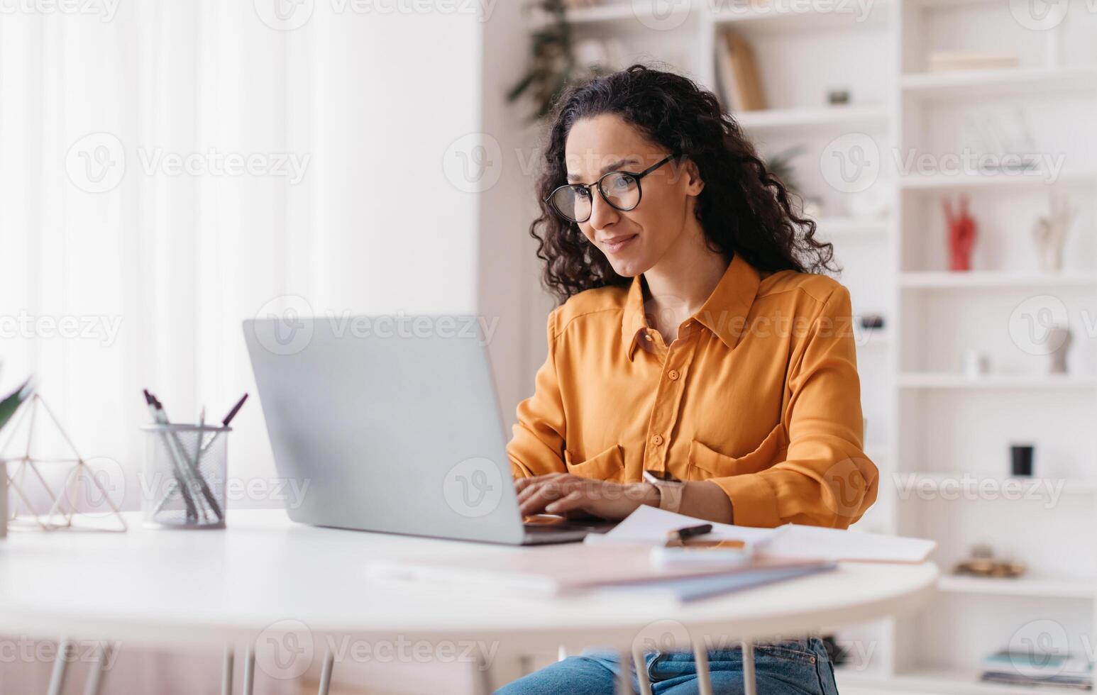 Middle Eastern Lady Using Laptop Working Online Wearing Eyewear Sitting At Workplace In Modern Office. Remote Job, Technology And Career Profession Concept. Side View photo