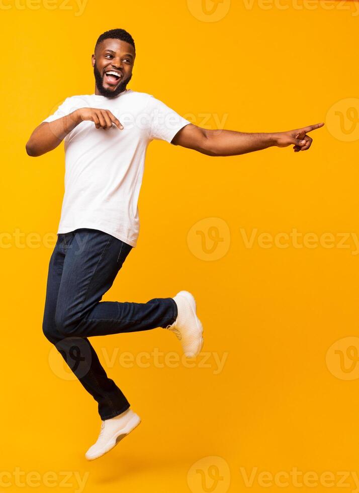 Joyful black millennial guy jumping on air and pointing aside, free space photo