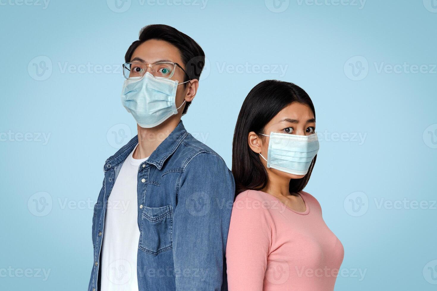 Afraid Asian Couple In Protective Medical Masks Standing In Studio Over Light Background, Panicking About Coronavirus Epidemy, Empty Space photo