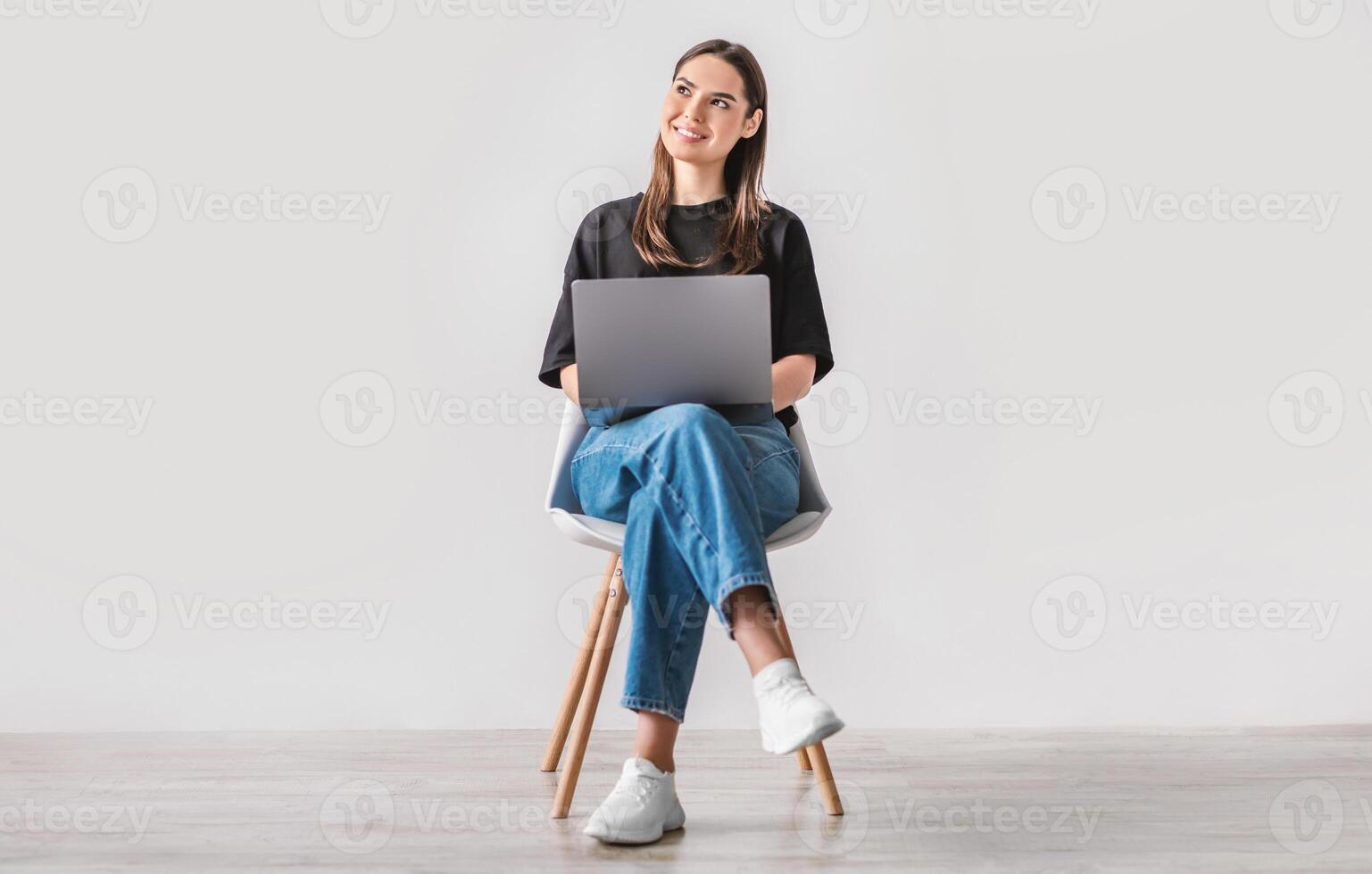 Beautiful young woman sitting on chair, working on laptop against white studio wall, full length. Dreamy millennial lady using portable pc for work, entertainment or online studies photo