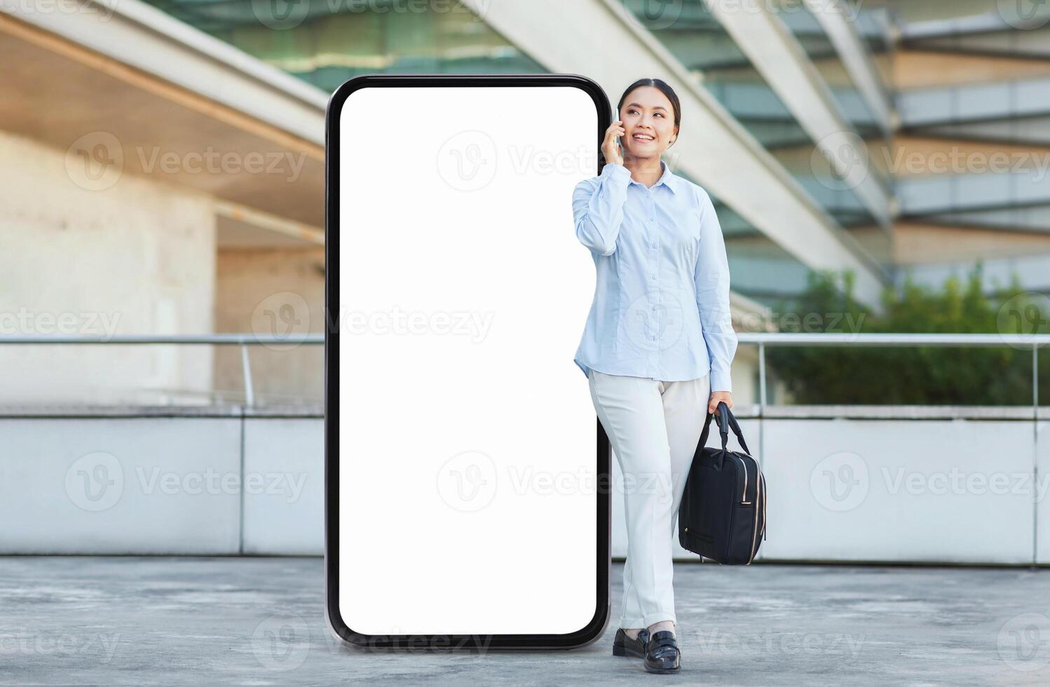 Asian woman smiles while talking on a phone, standing next to a large blank phone screen in an outdoor urban setting, dressed in casual attire, ready for her day ahead. photo