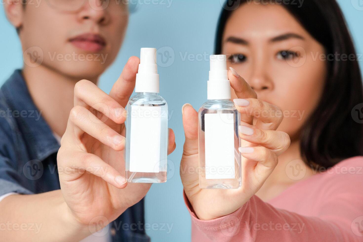 Hand sanitizer. Young asian couple demonstrating bottles with antibacterial spray, standing on light background in studio, selective focus, mockup photo