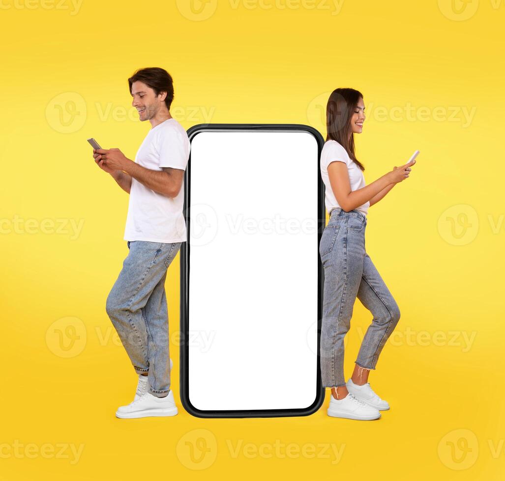 Great App. Excited Couple Leaning On Big Smartphone With Blank White Screen Using Cell Phones, Cheerful Guy And Lady Chatting On Social Media, Standing On Yellow Orange Background, Mock Up, Full Body photo