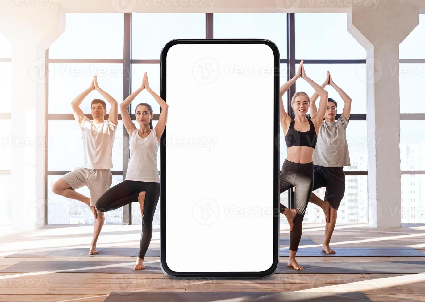 A group of four individuals performs yoga poses in a bright, modern studio. The large windows let in natural light, and a smartphone stands in the center, inviting a digital connection to wellness. photo