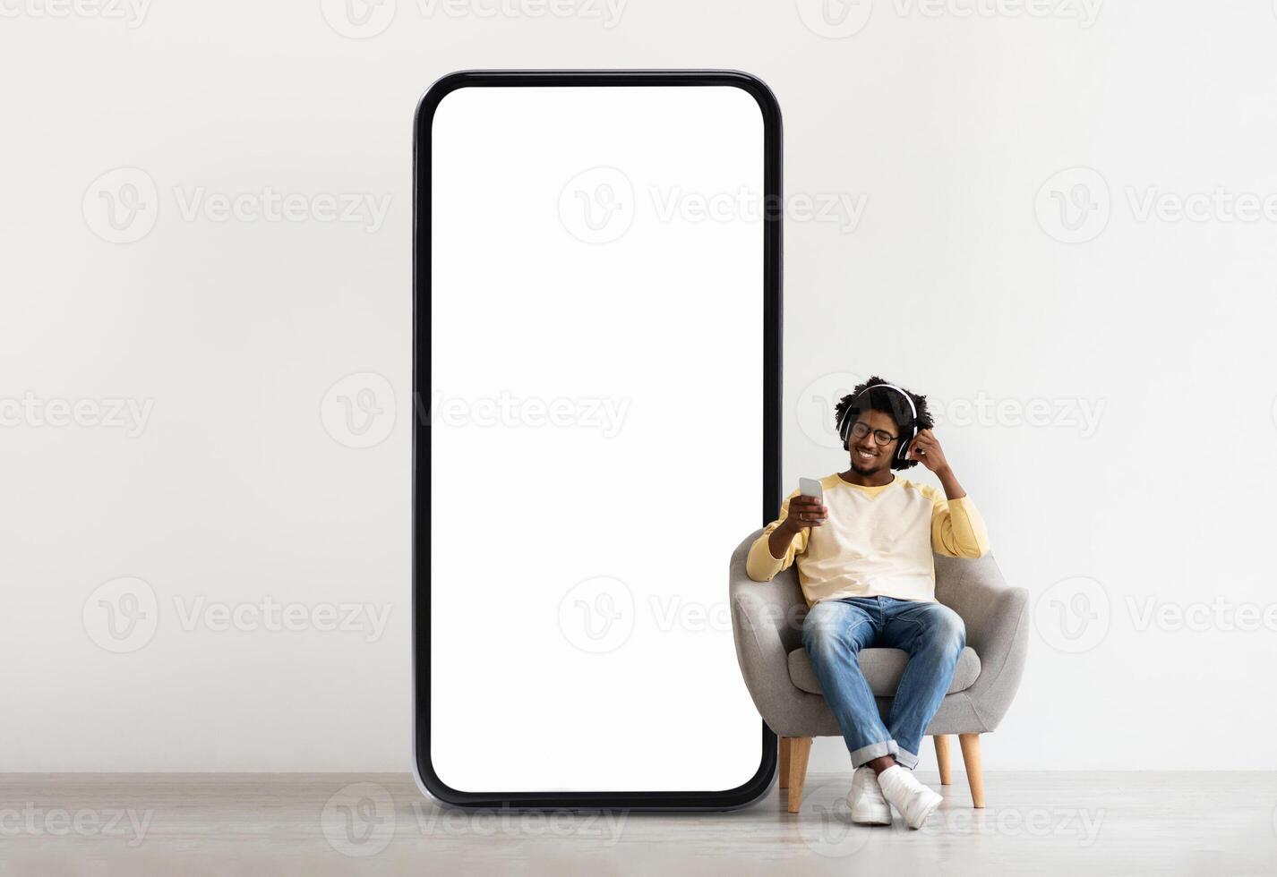 A young black man sits comfortably in a chair, enjoying his smartphone as he listens to music. Next to him stands a large blank screen, creating a minimalist atmosphere. photo