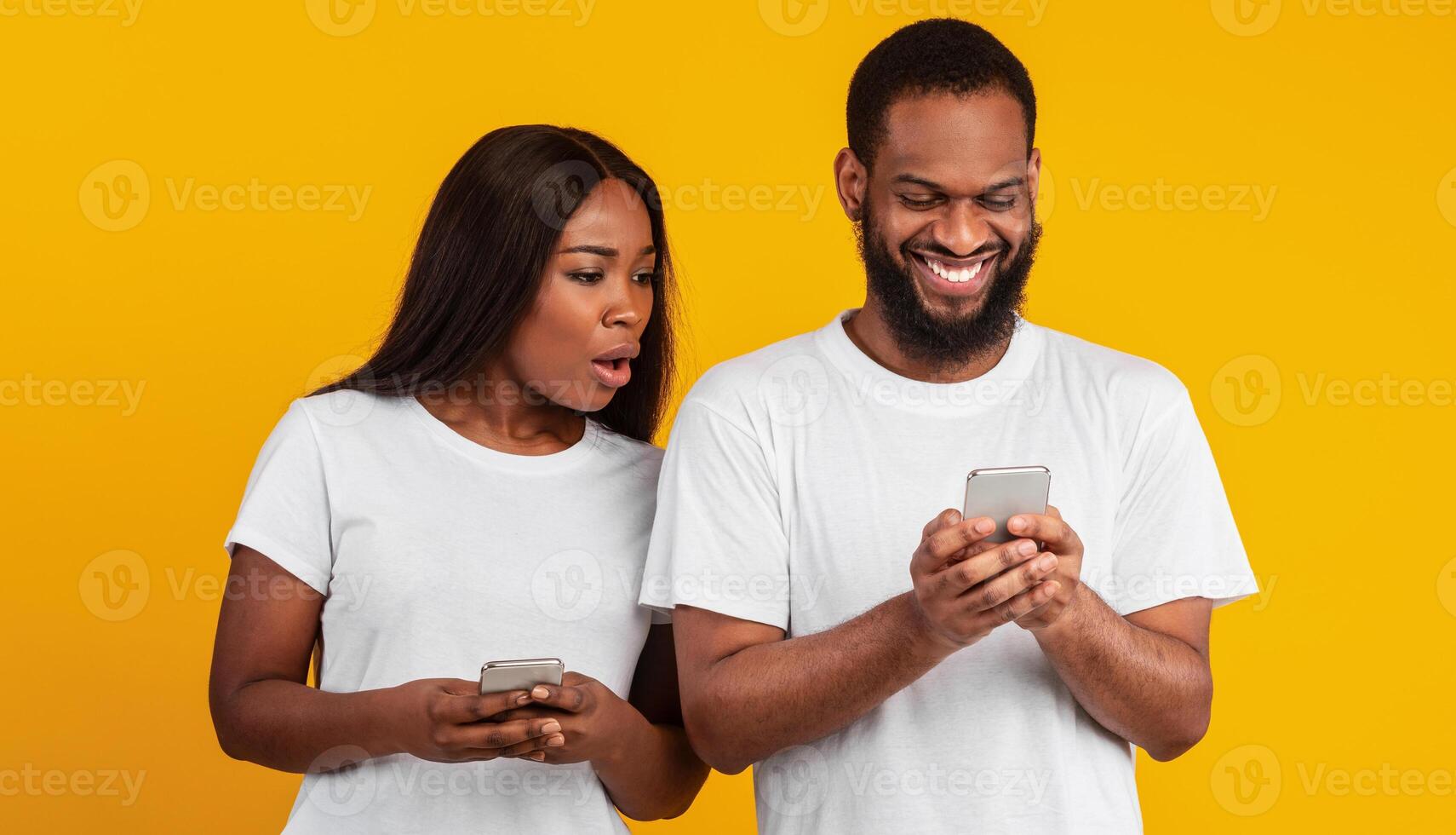 Portrait of shocked African American woman spying on her smiling boyfriend who using mobile phone, texting sms or scrolling social media news feed. Black couple standing over yellow studio background photo