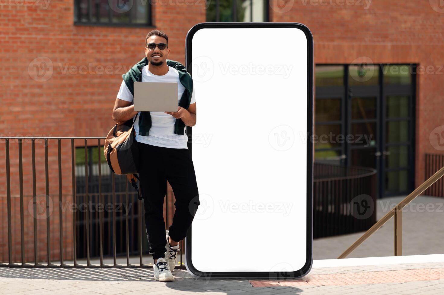 Black man stands confidently with a laptop in one hand beside a blank, phone-shaped display near a modern building. He is casually dressed, ready for work or study in an urban environment. photo
