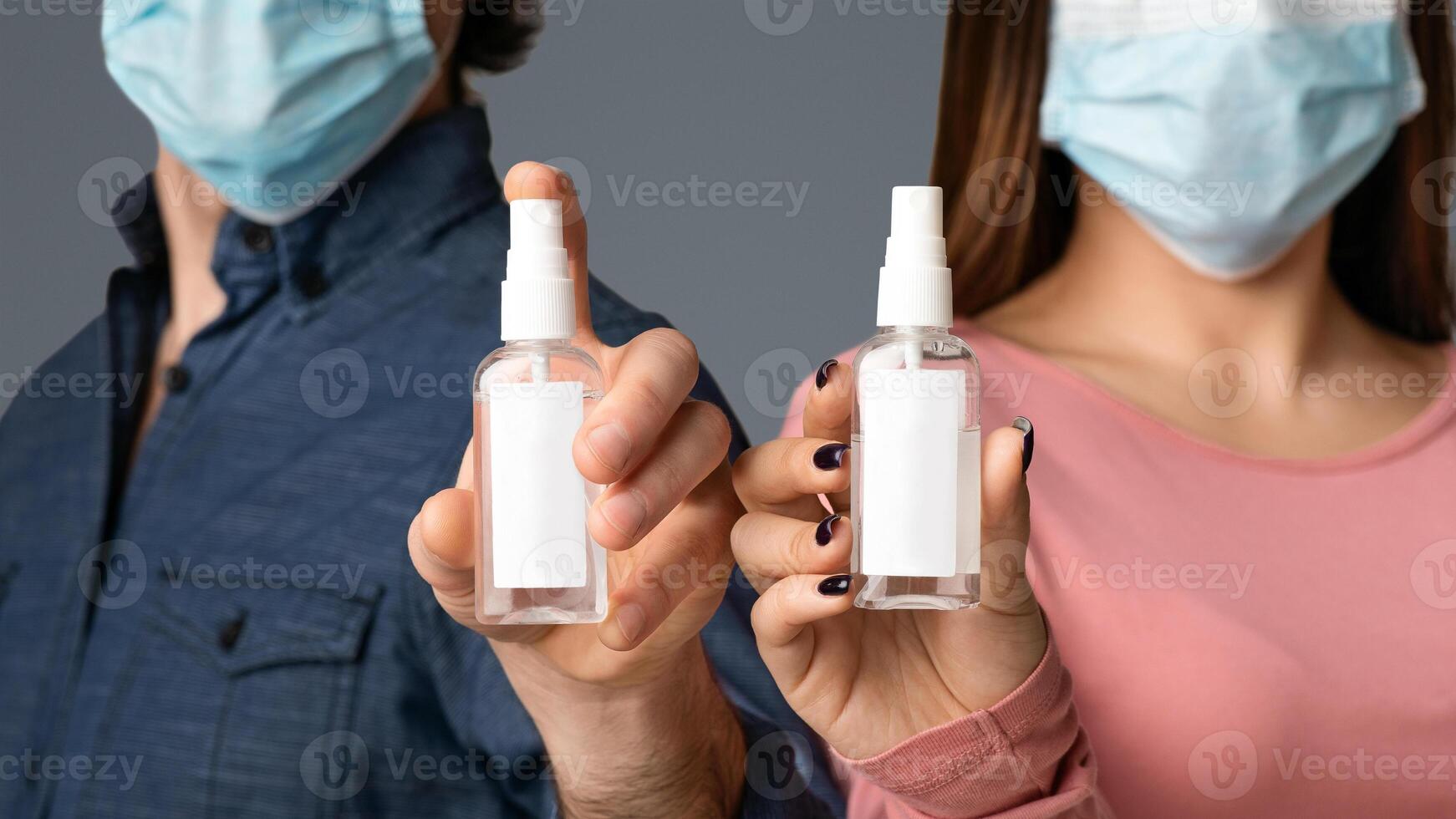 Bottles Of Antibacterial Disinfectant Sprays With Blank Sticker Labels In Hands Of Young Couple Wearing Medical Masks, Light Background, Mockup photo