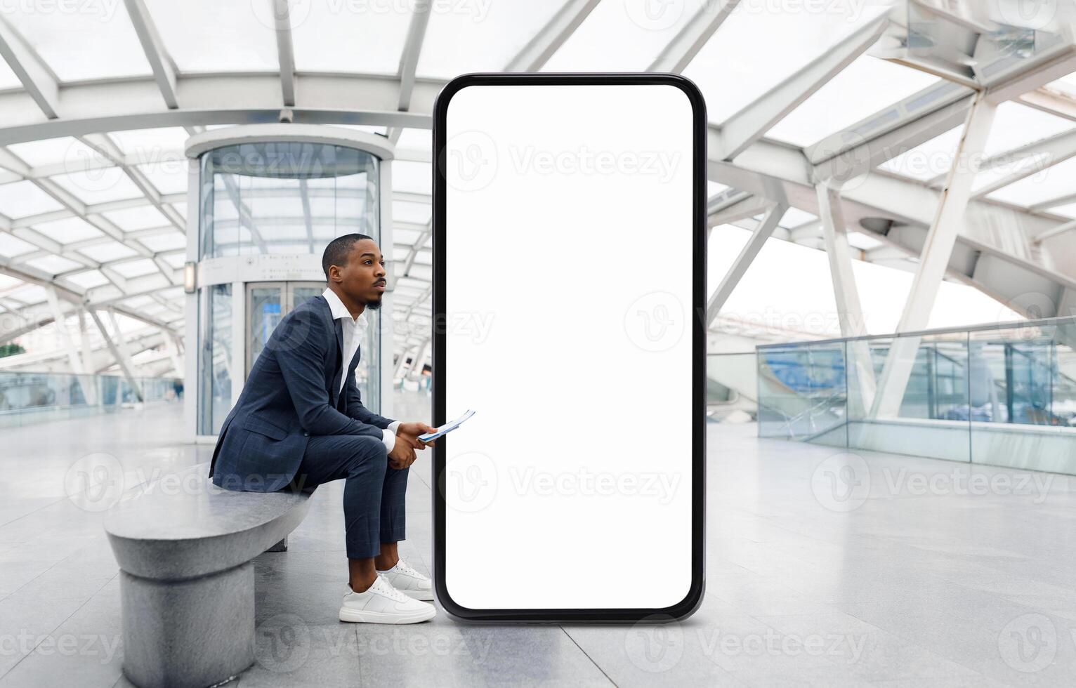 African American man in a suit is seated on a bench in a contemporary architectural space, appearing thoughtful as he waits next to a large, empty digital display. photo