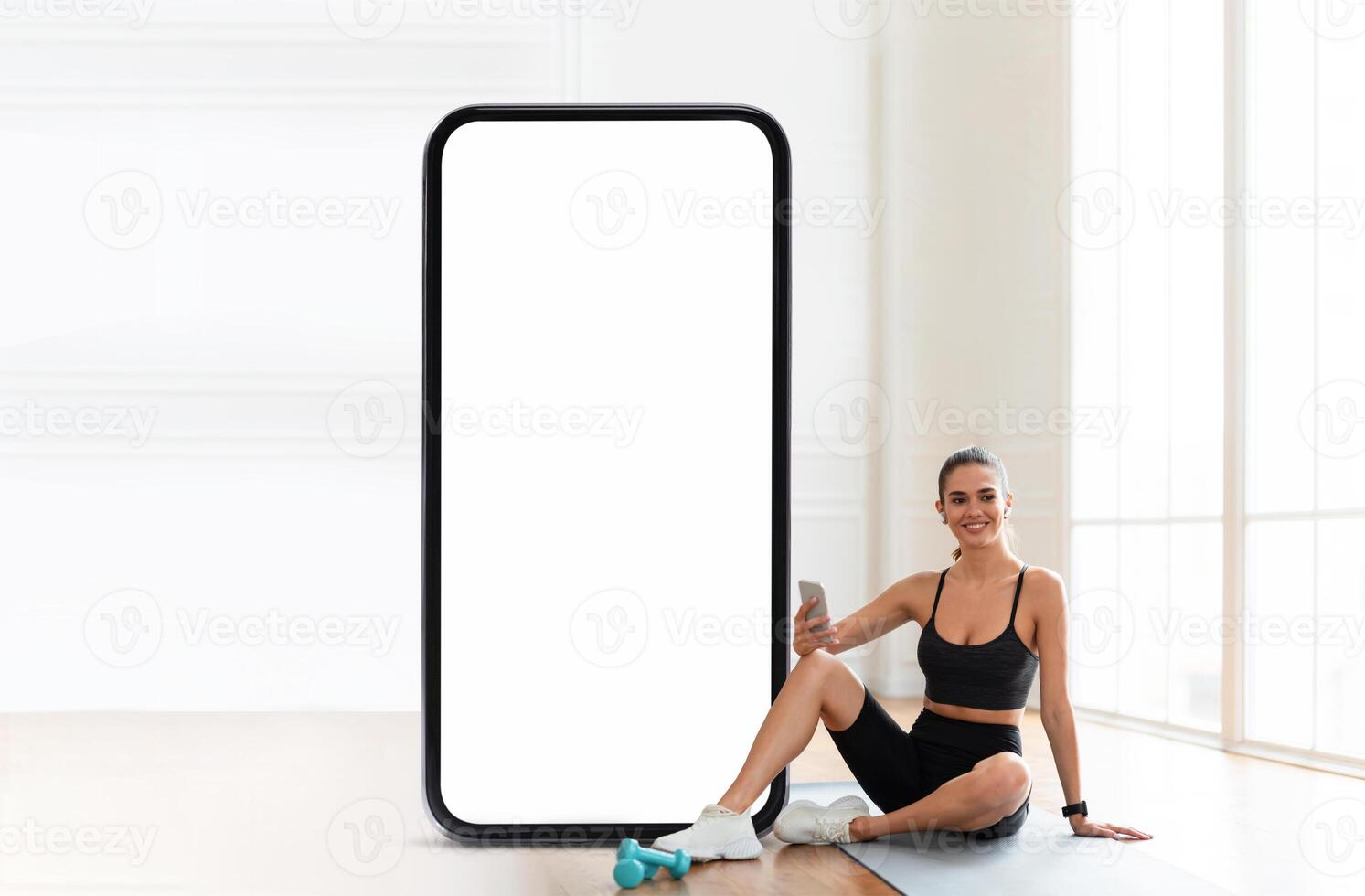 A woman in workout attire sits on the floor next to a large blank display, holding a phone while resting after exercising, with bright ambient light and a minimalistic background. photo