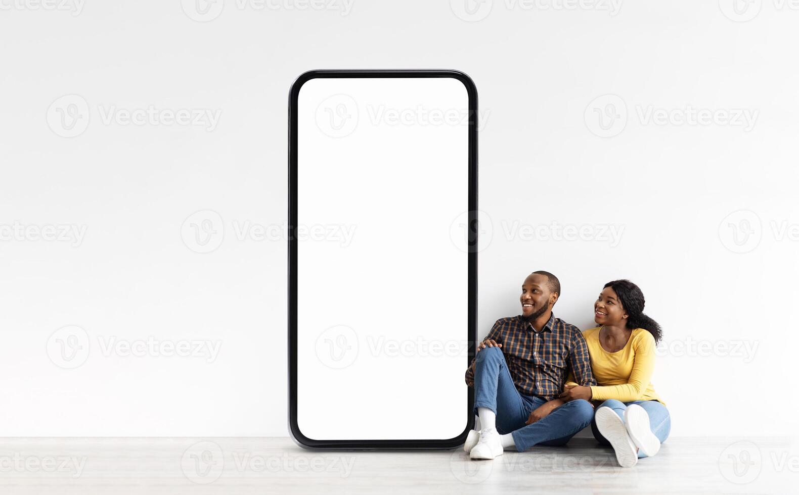 Black couple sits side by side on the floor in a modern indoor setting, smiling and laughing while resting against a large blank screen. They appear relaxed and content in each other's company. photo
