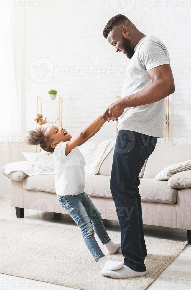 Playful black daughter staying on dad feet, learning dancing at home photo
