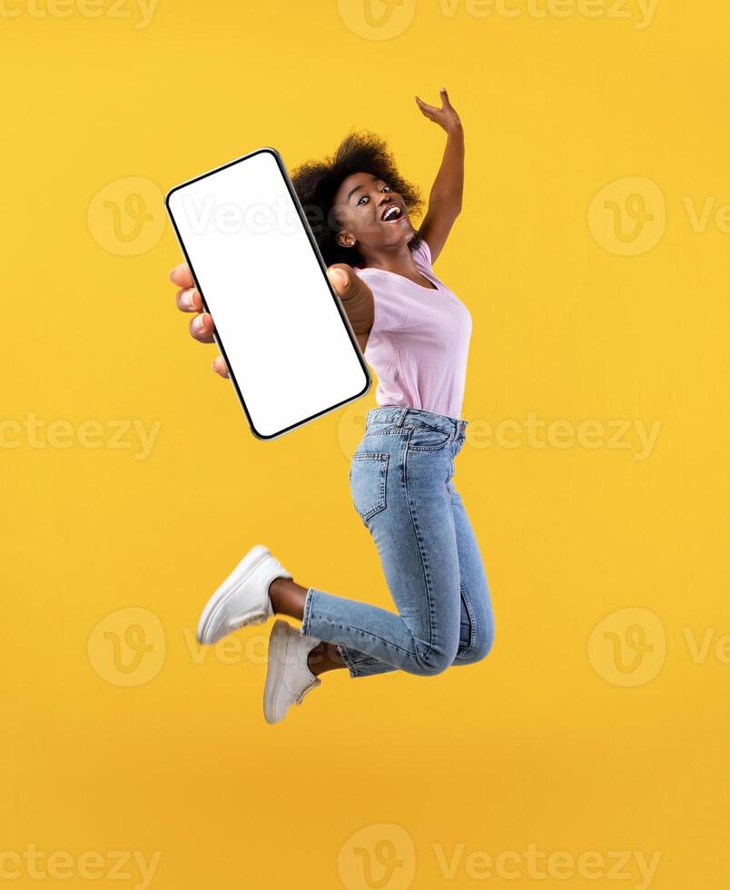Overjoyed african american young lady showing empty smartphone screen while jumping up over yellow studio background, collage, full size photo. Happy black woman recommending new mobile app photo