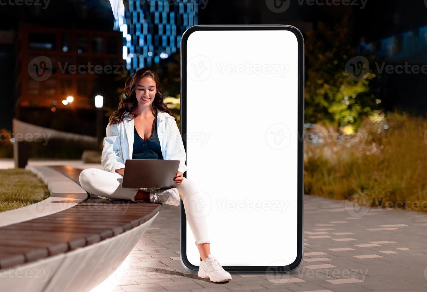 A young woman sits comfortably on a park bench, using her laptop in front of a large blank screen at night. The urban surroundings are softly illuminated by nearby lights. photo