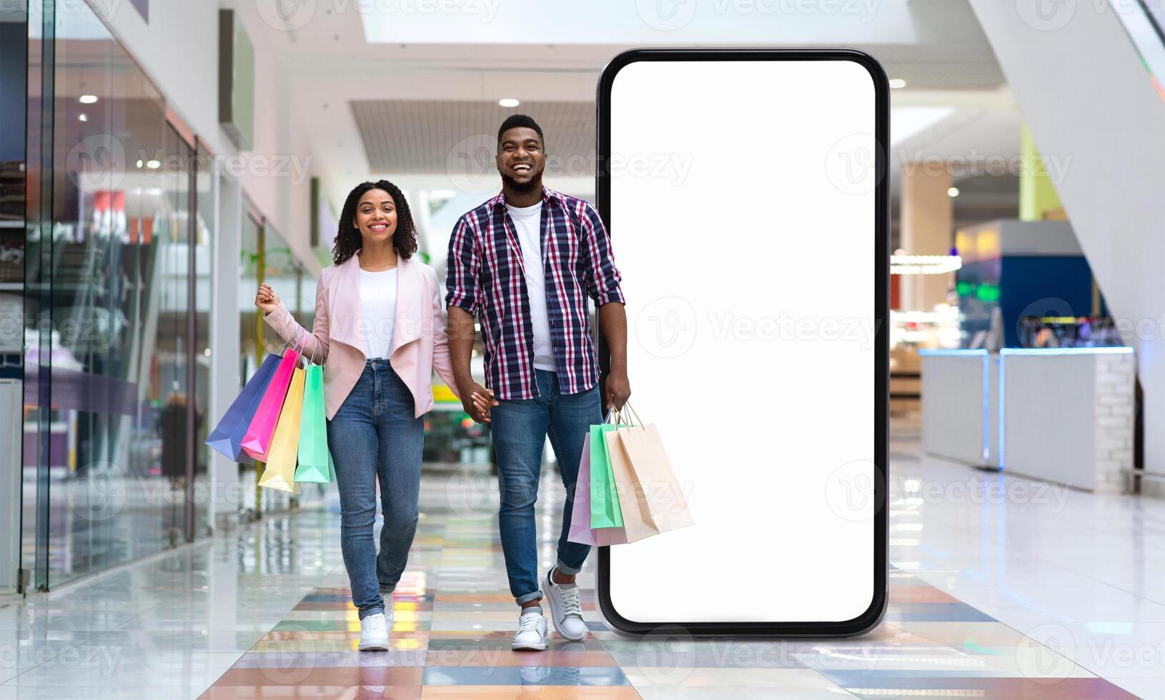 A joyful black couple strolls through a modern shopping mall, holding colorful shopping bags. They walk beside a large, empty display screen, creating a vibrant atmosphere. photo
