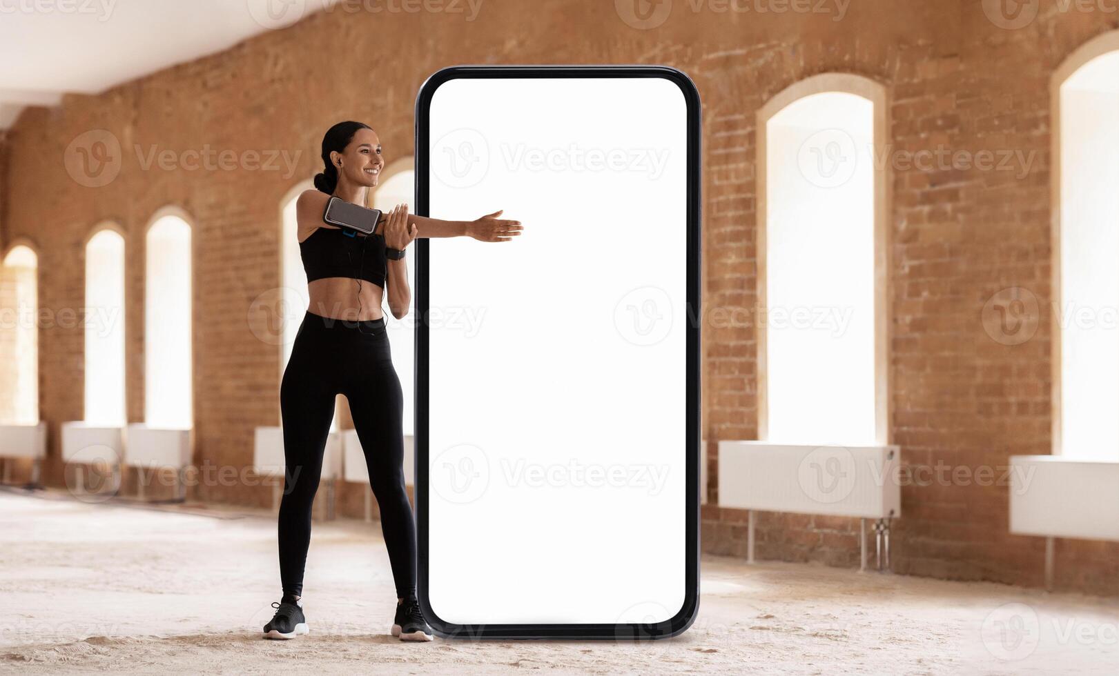 A woman in athletic gear is stretching while standing next to a large, blank screen in a spacious, well-lit indoor setting. The atmosphere is energetic and focused on fitness. photo