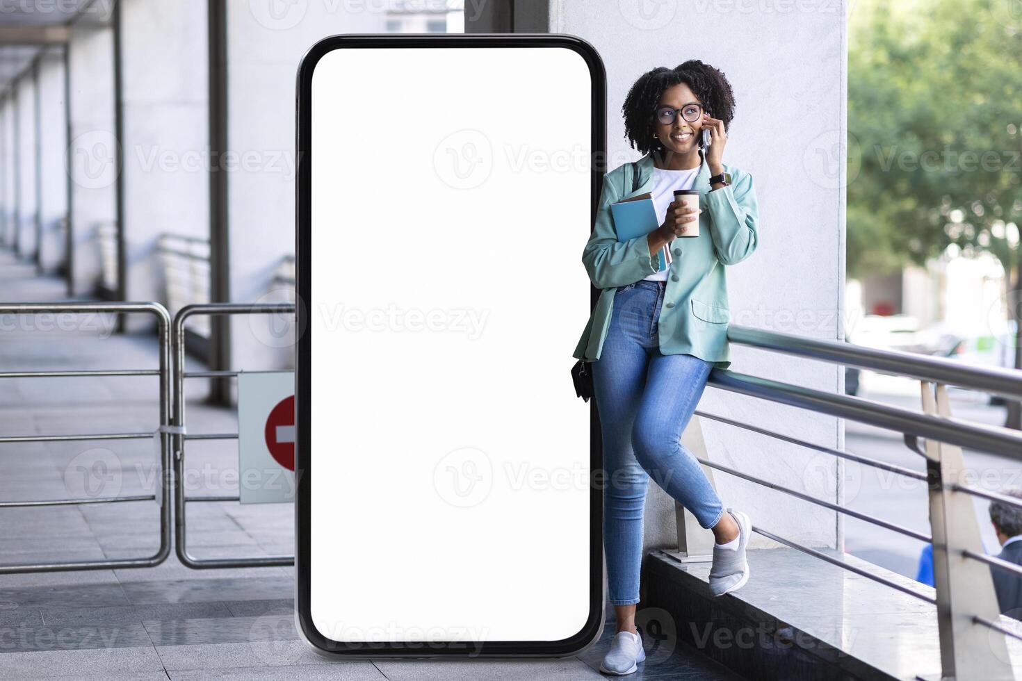 In an outdoor setting, a young black woman casually chats on her phone while leaning against a large blank screen, holding a drink and a book in her other hand, enjoying her time. photo