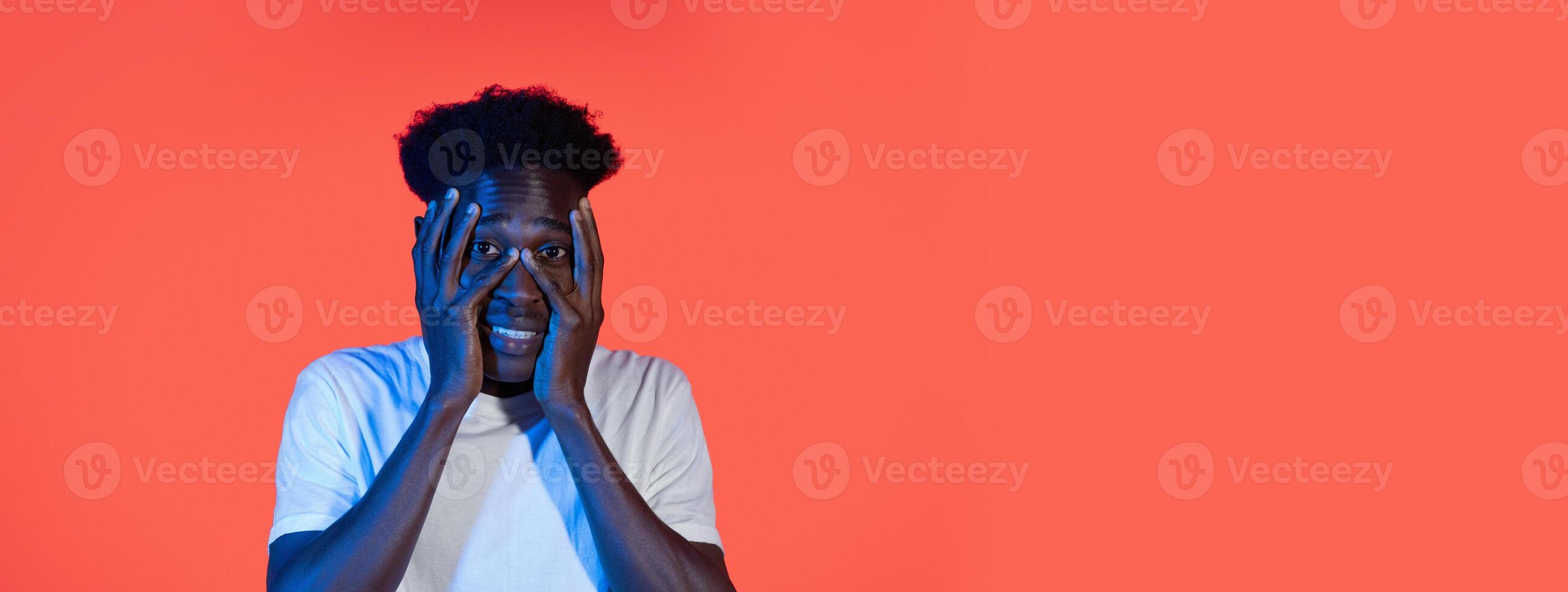 A young black man stands in front of a bright orange backdrop, showcasing a surprised expression with hands on his face. His casual attire contrasts with the vivid color around him. photo