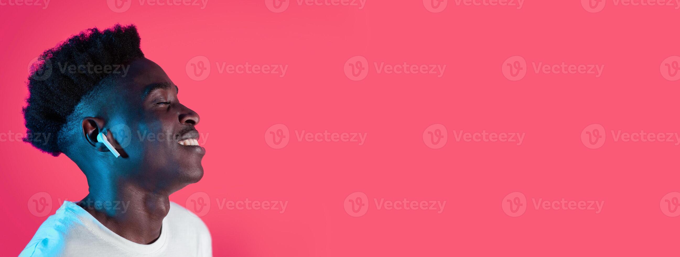 A young black man enjoys music through wireless earbuds, smiling with his eyes closed. The vibrant pink backdrop enhances the joyful atmosphere, capturing a moment of relaxation and enjoyment. photo