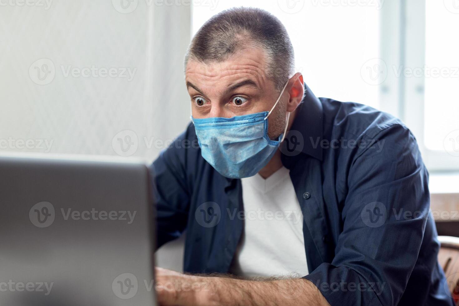 Coronavirus And Crisis. Shocked Man Working On Laptop Computer Wearing Protective Mask Sitting At Desk At Home During Covid-19 Outbreak. photo