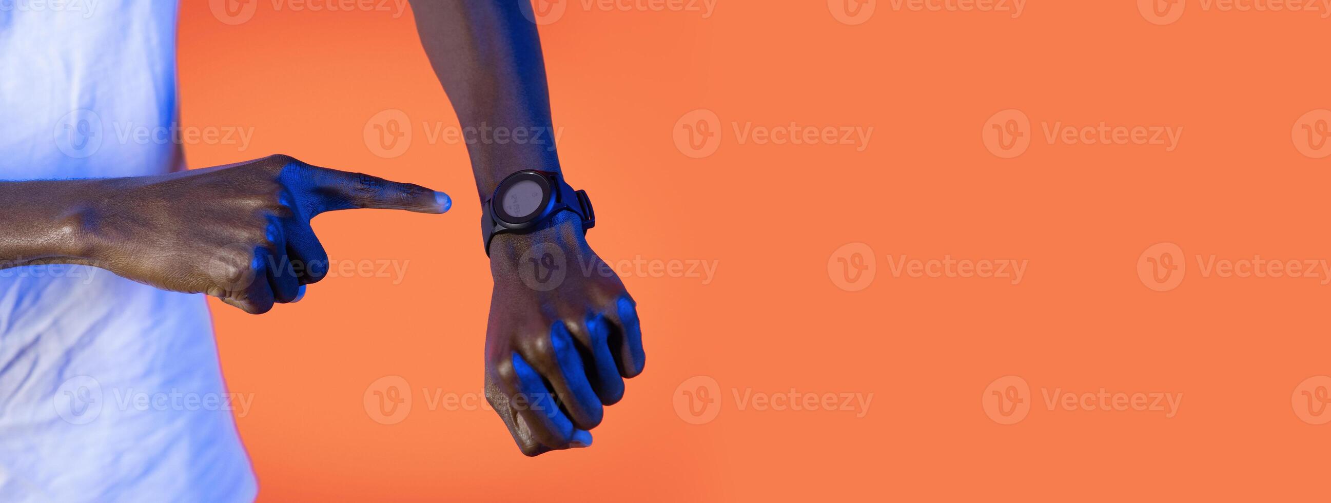 Black guy pointing at his wristwatch, showcasing its features. The bright orange background creates a vibrant contrast that highlights the watch's details, cropped photo