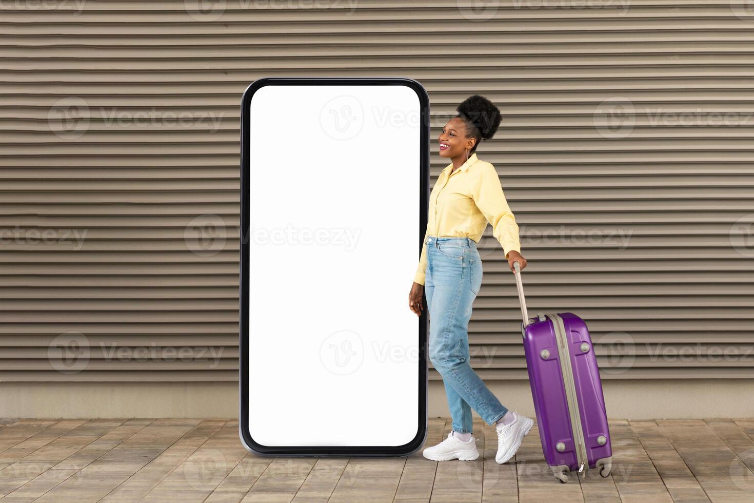 A black woman stands next to a large blank advertising board, holding a purple suitcase while smiling. She is dressed casually, reflecting a contemporary urban lifestyle. photo