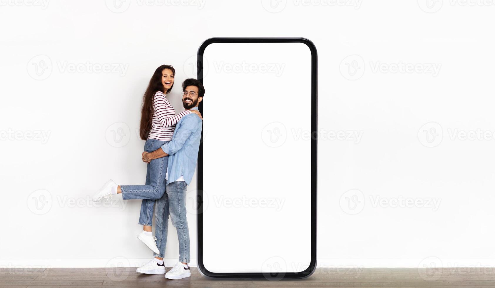 Indian couple stands next to an oversized blank phone display in a well-lit indoor setting, with one partner playfully lifted off the ground, showcasing a fun and dynamic moment. photo