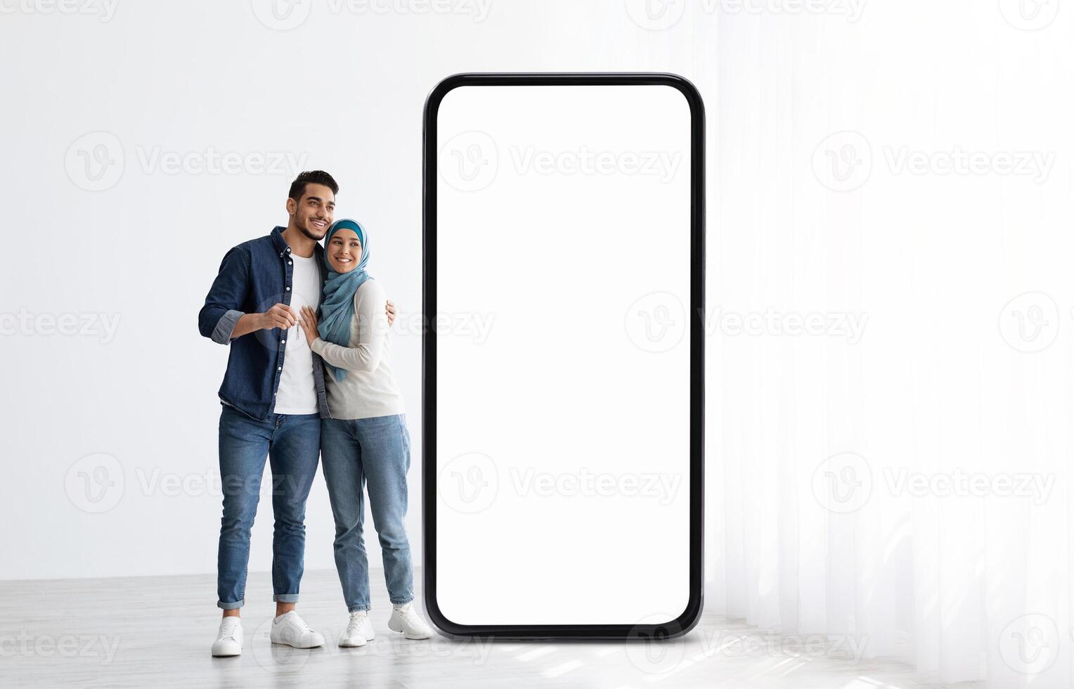 A young Muslim couple stands side by side, beaming with joy while leaning against a large, blank smartphone screen in a bright indoor space filled with natural light. photo