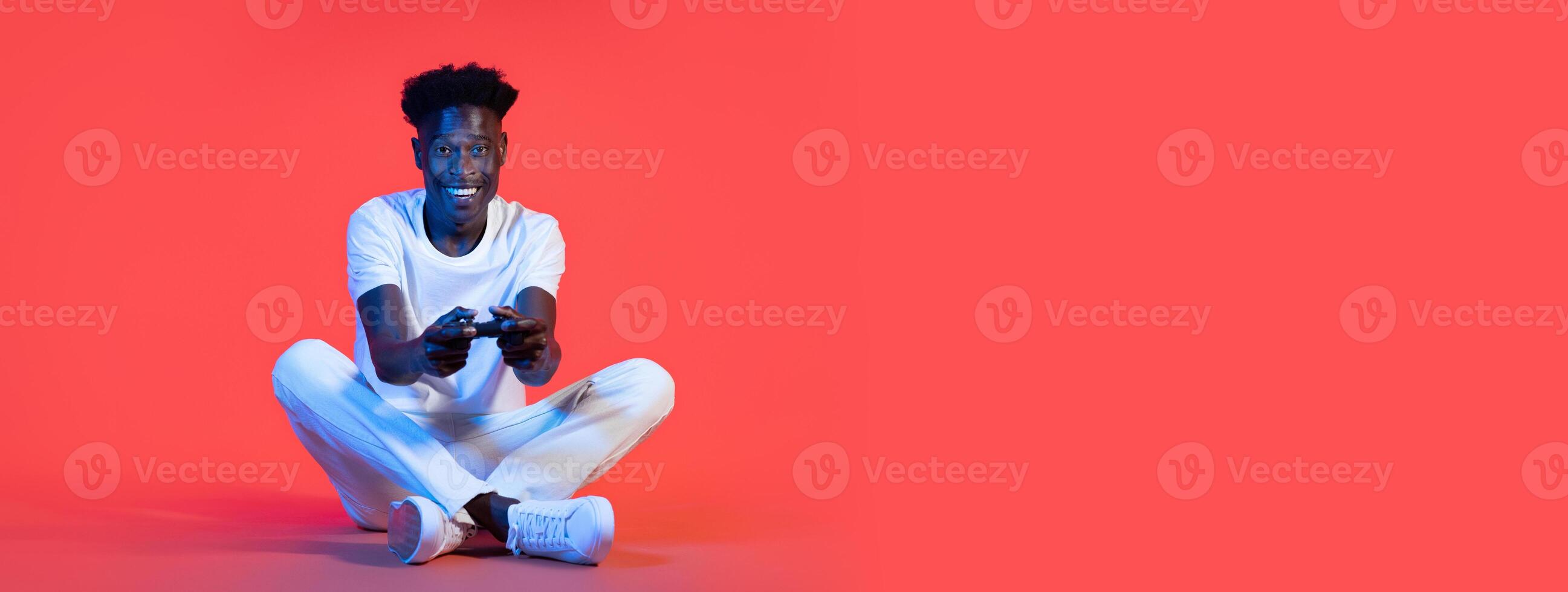 African American young man is comfortably seated on the floor, focused on a gaming controller in his hands. The vibrant background adds energy to the engaging activity of gaming. photo