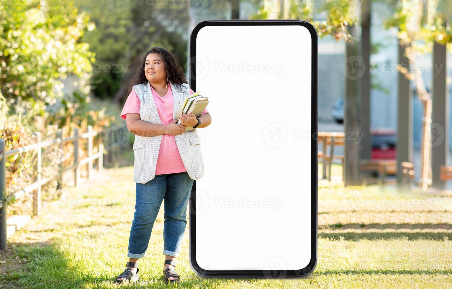 A young woman holds books while standing next to a large blank sign in a park. The scene is bright and sunny, surrounded by greenery and trees. photo