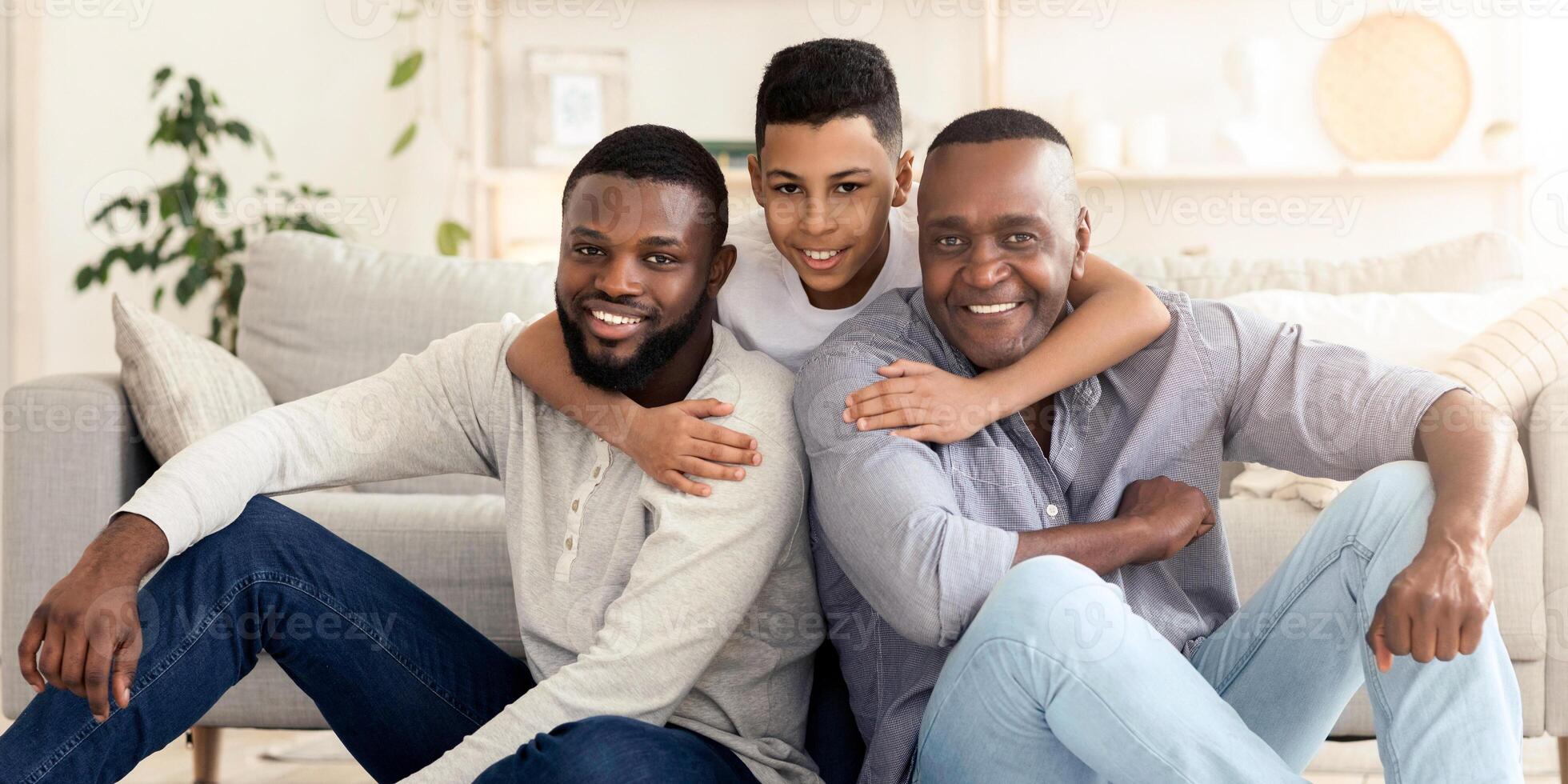 Happy preteen black boy hugging his father and grandpa, sitting on floor at home, looking at camera photo