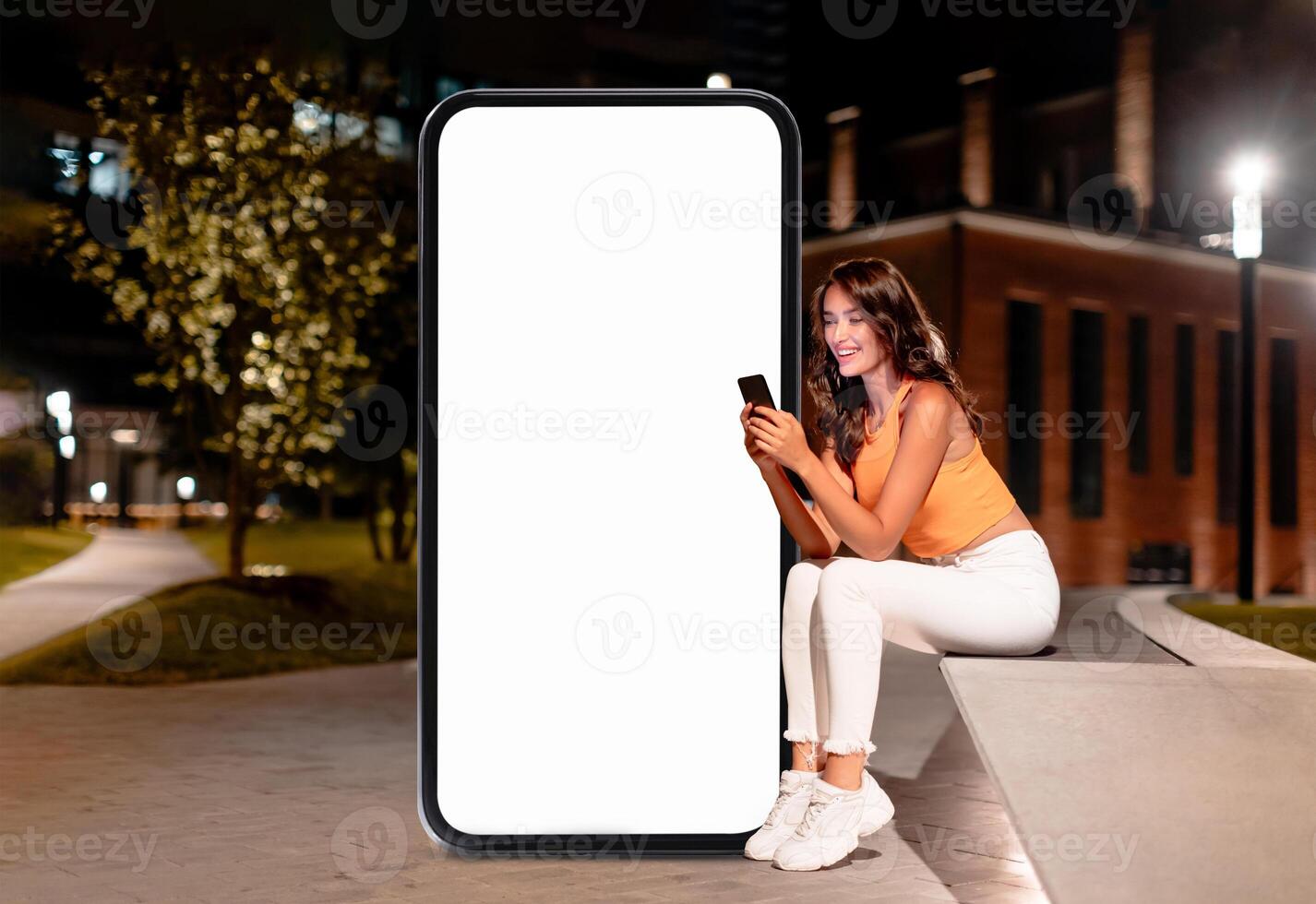 A young woman sits comfortably on a bench, engaged with her smartphone. A large blank screen stands beside her, illuminated by nearby lights in a tranquil outdoor setting. photo