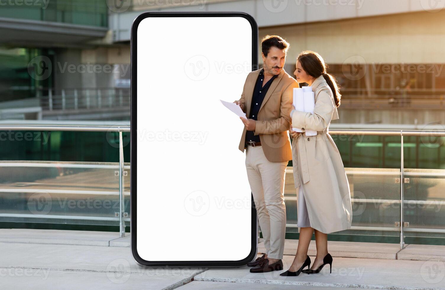 A business couple stands outdoors near a large blank screen, engaging in a conversation while reviewing documents. The sun sets in the background, creating a warm atmosphere. photo