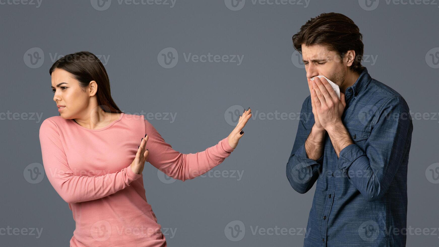 Social Distancing. Young woman disgusted by her sick boyfriend with runny nose, afraid of coronavirus spread, light studio background with free space photo