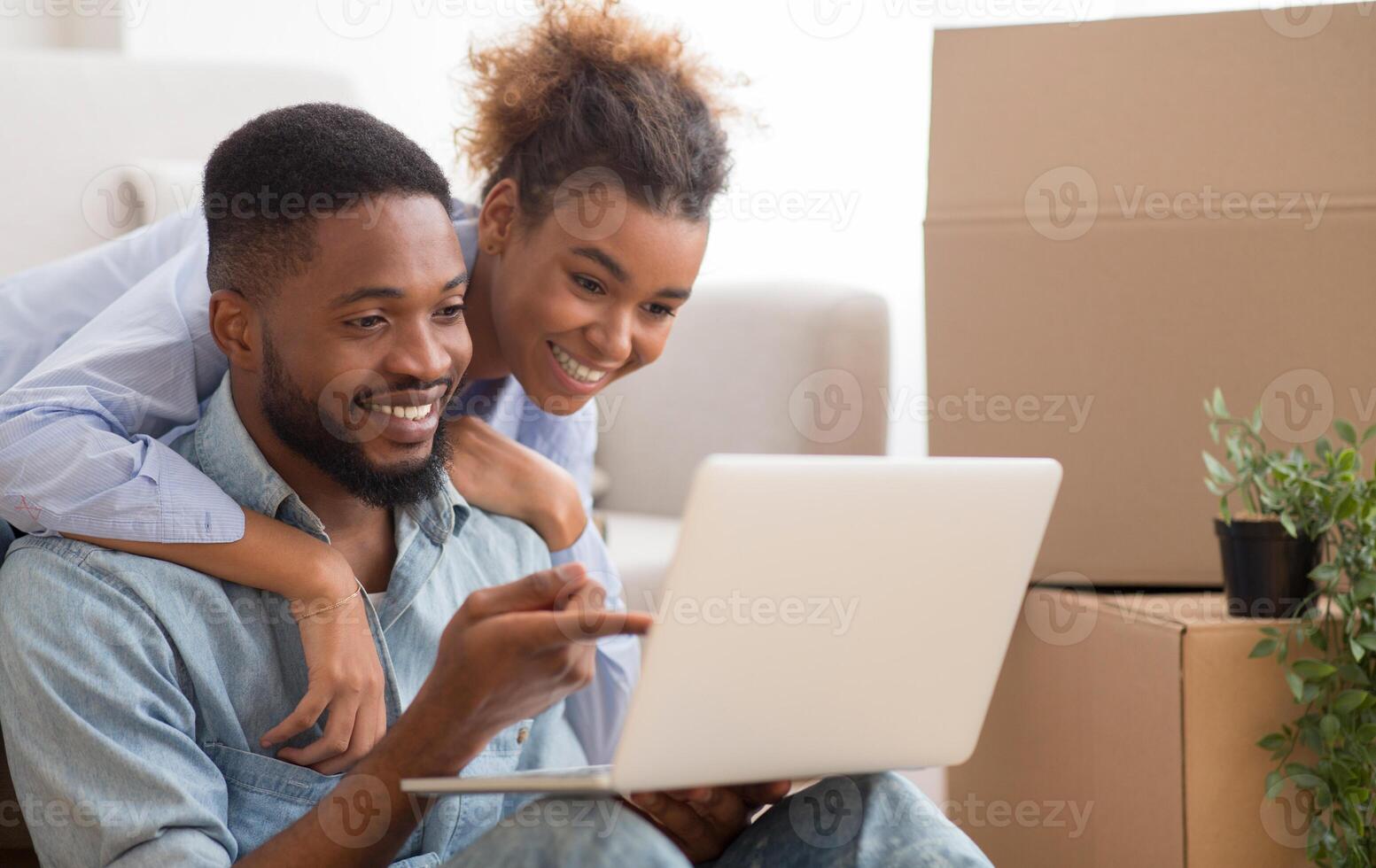 Smiling Black Spouses Using Laptop Sitting On Sofa Among Packed Boxes In New Home. Moving House Concept. Selective Focus photo