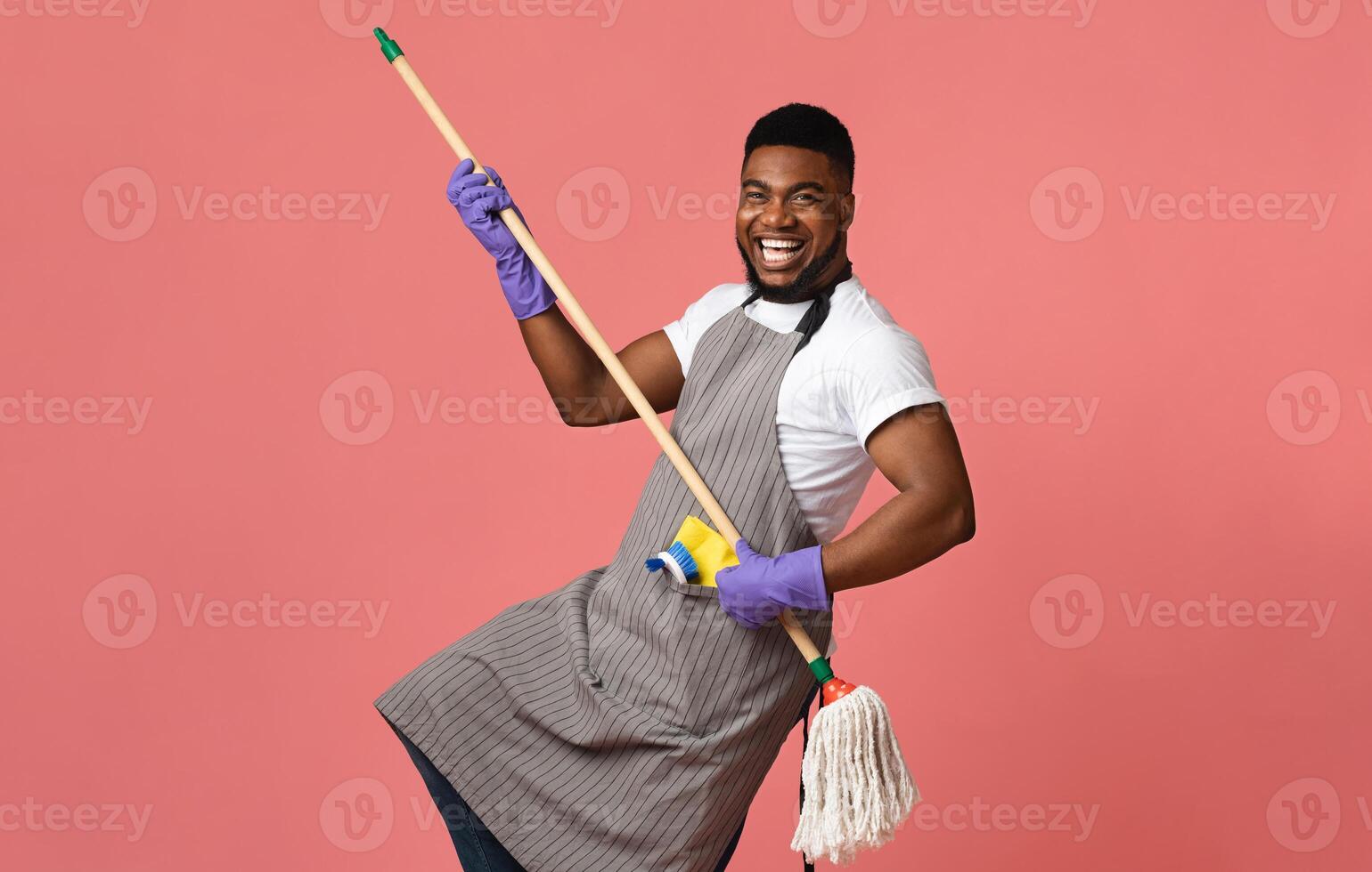 Portrait Of Funny Black Man In Apron With Mop In Hands Posing Over Pink Studio Background, Using Handle As Guitar, Fooling With Household Tool photo