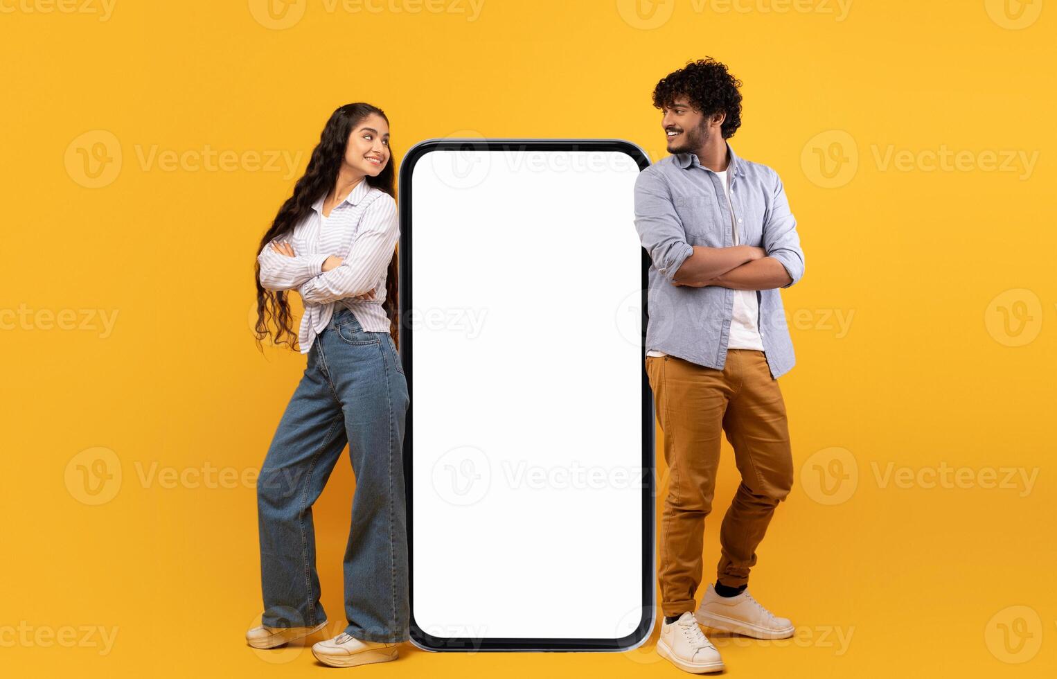 Mobile offer. Millennial indian couple leaning on giant smartphone with mockup, advertising or promoting app or website over yellow studio background photo