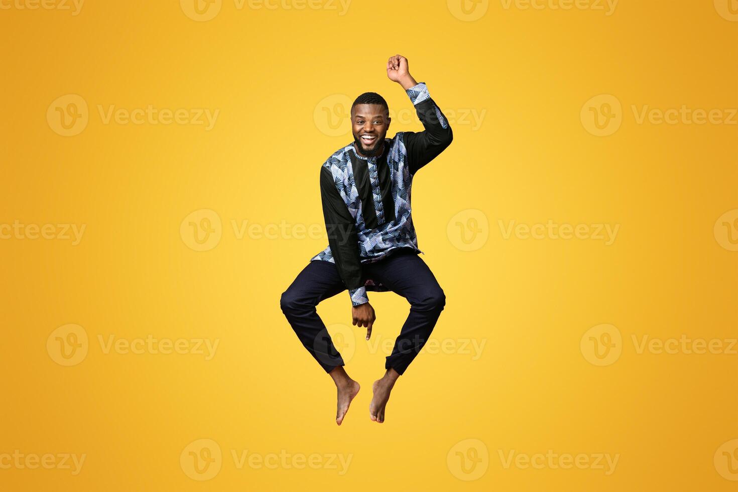 Funny black man in african costume jumping in the air on pink studio background. Smiling african american guy having fun, wearing traditional clothes, full size photo. Traditions and culture concept photo