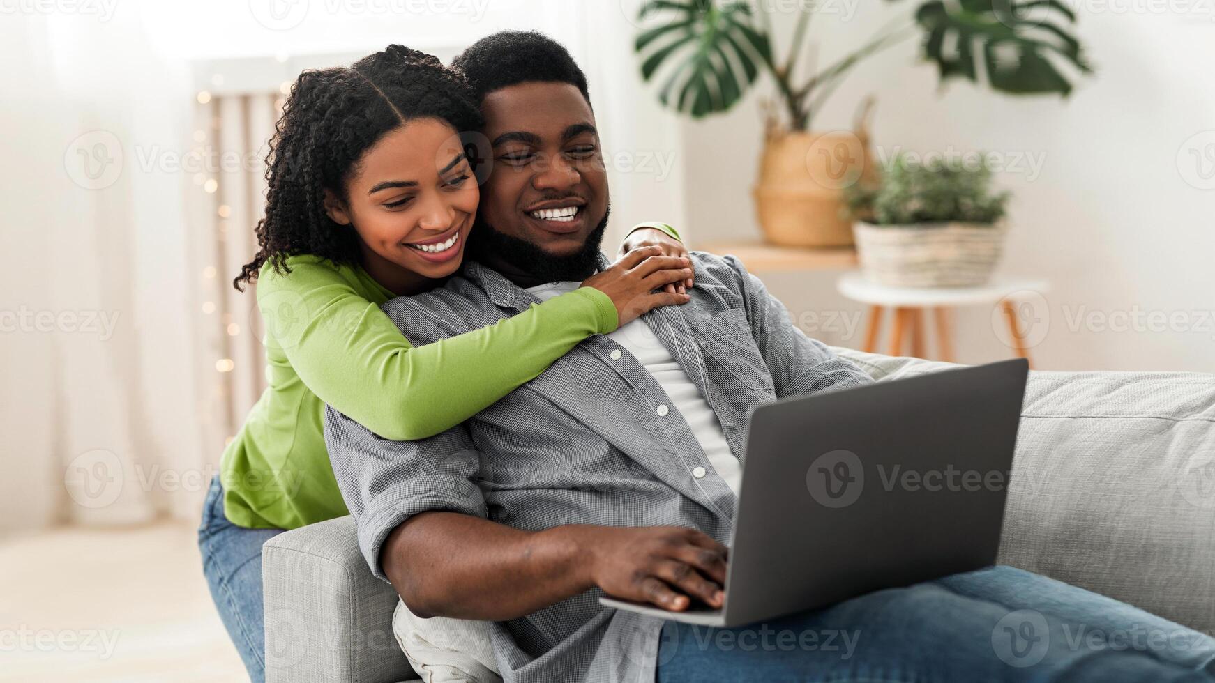 Cheerful black couple web surfing on laptop together, sitting on sofa at home, relaxing and hugging, copy space photo