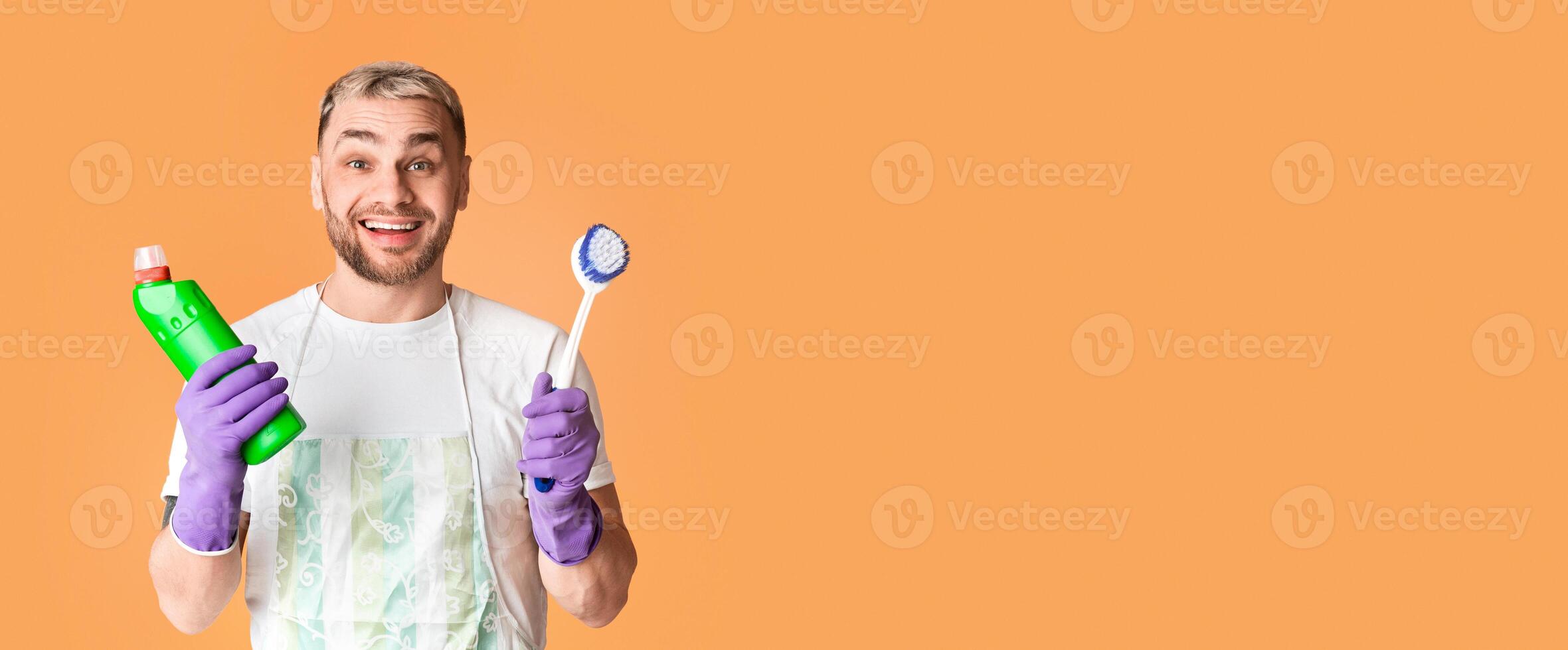 Smiling emotional guy in apron holds brush and cleaning supply on yellow background photo