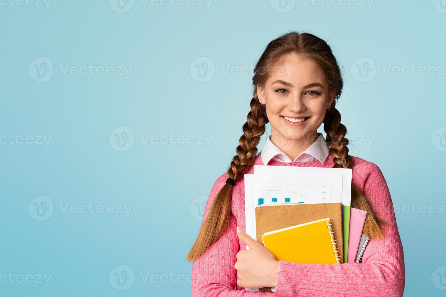 Study on college or school concept. Smiling teenage girl holds notebooks and charts and looks at empty space, isolated on blue background, studio shot photo