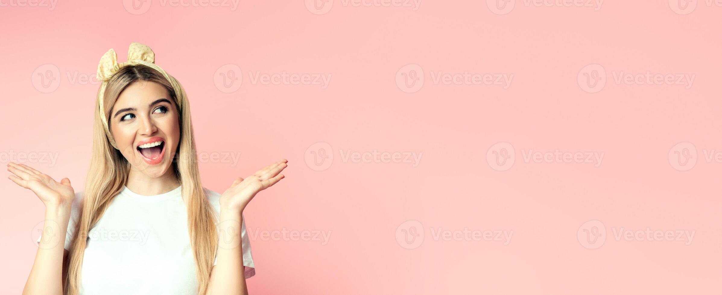 Place For Ad. Surprised happy blonde girl looking up to the side at copyspace in excitement, spreading arms, pink wall photo