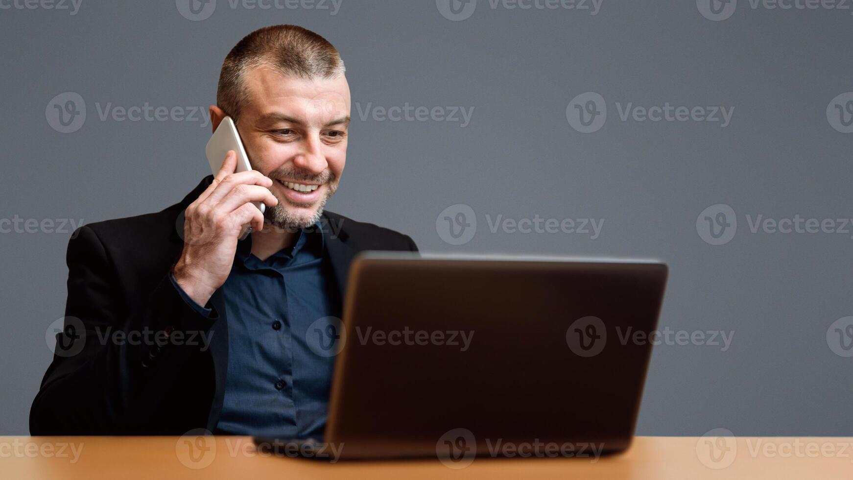 Businessman Working On Laptop Talking On Cellphone Sitting Over Gray Background. Studio Shot, Selective Focus photo