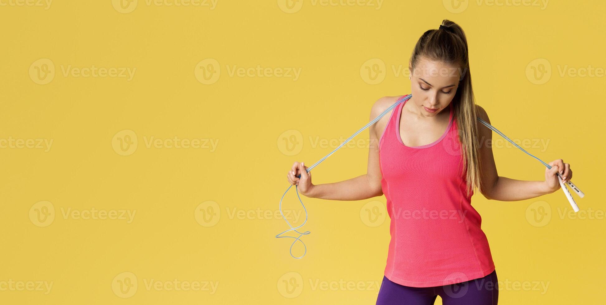 Young woman with jump rope smiling looking at her fit body, posing on yellow studio background with free space photo