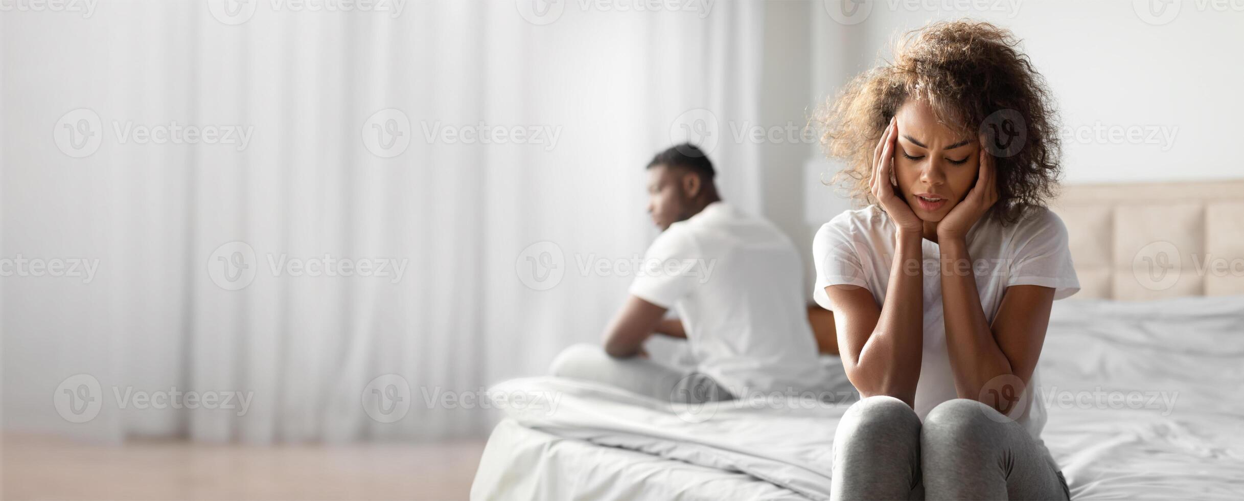 A black woman sits on the edge of a bed, holding her head in distress, while a man sits apart from her, symbolizing emotional distance and communication issues in their relationship. photo