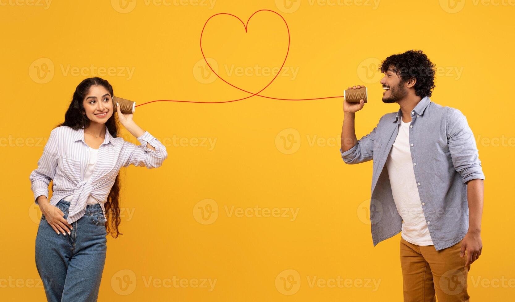 Love message. Loving indian man telling romantic words to girlfriend through tin can phone with heart shaped string, standing over yellow background, creative collage photo