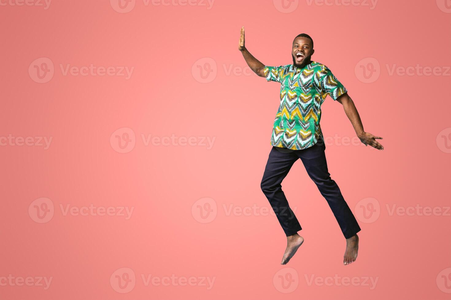 Smiling african american guy having fun, wearing traditional clothes and jumping up in the air over blue studio background. Joyful black man in bright african costume posing at camera photo