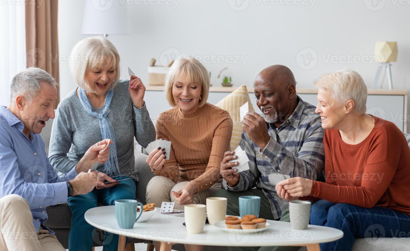 Multiethnic group of happy senior friends men and women in casual sitting on couch, having conversation and playing cards while drinking tea with cookies, chilling together at home photo