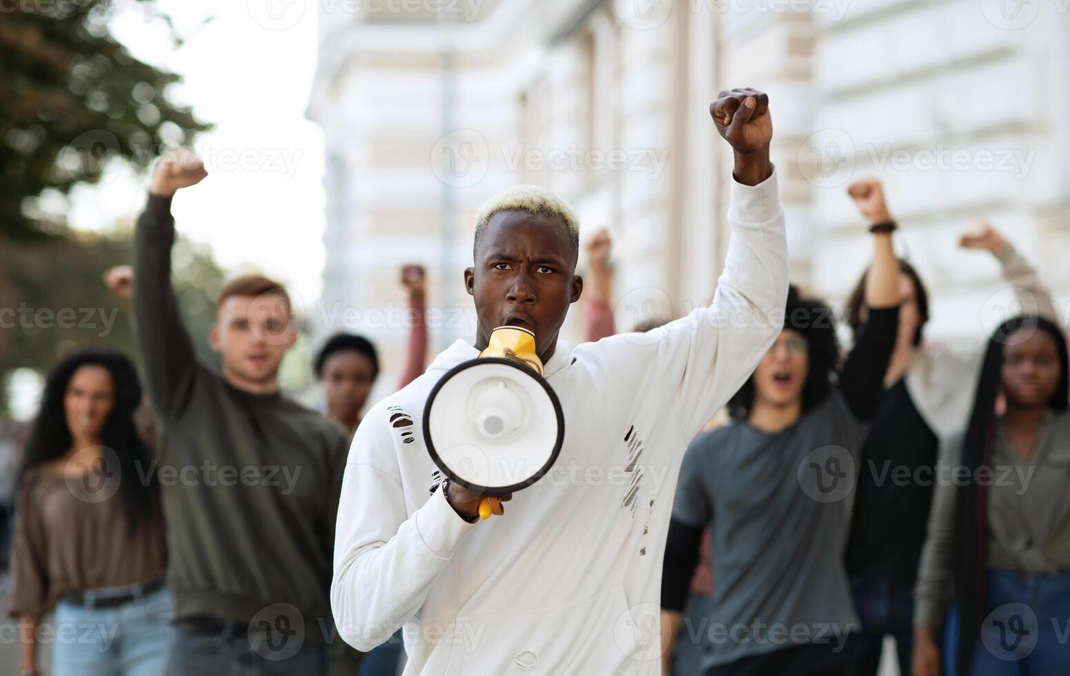 African american man with megaphone leading international group of protestors on the street, raising fist up, chanting slogans. Leader of strikers with loudspeaker yelling demands to government photo